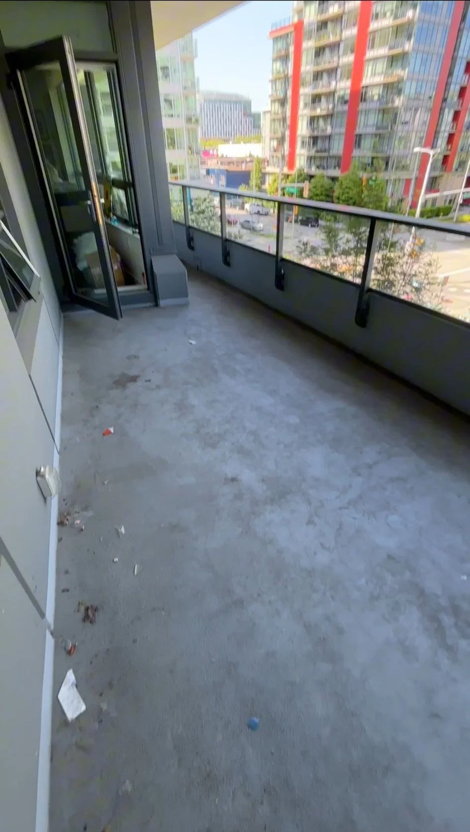 Empty apartment balcony with concrete floor, glass railing, and city view of modern buildings.