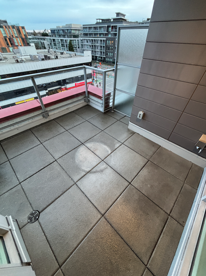 Empty city balcony with wet tiled floor, glass railing, and view of modern buildings and a street below.