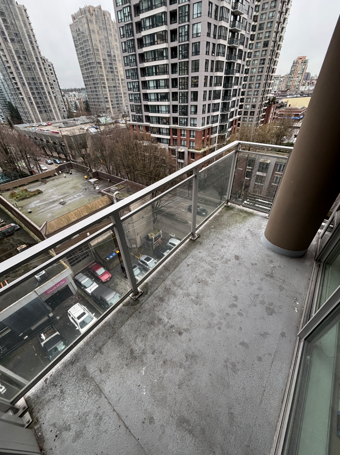 A small, partially damp balcony with concrete flooring, scattered leaves, and a glass railing overlooks a rainy, overcast street lined with trees.