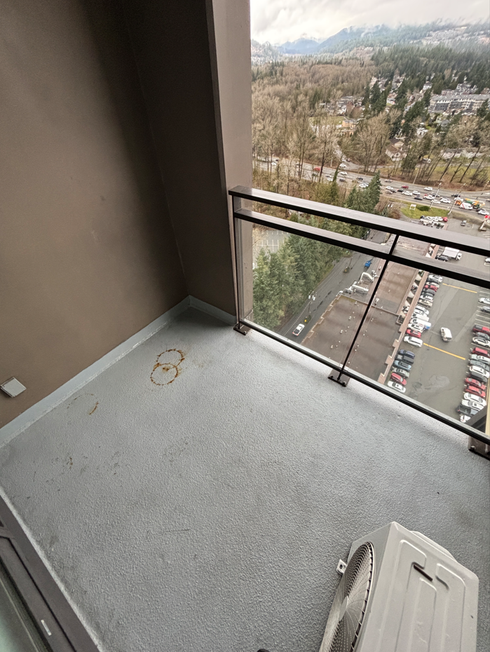 Small balcony with gray textured flooring and a metal railing, with stains on the floor. There is an air conditioning unit on the balcony. The view shows a parking lot below, trees, a road with cars, and distant mountains under cloudy skies.