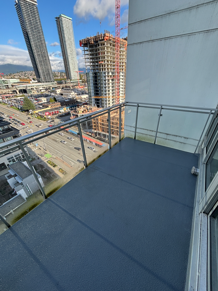 View from a high-rise balcony showing busy city streets, construction crane, and tall buildings under a blue sky with scattered clouds. Urban atmosphere.