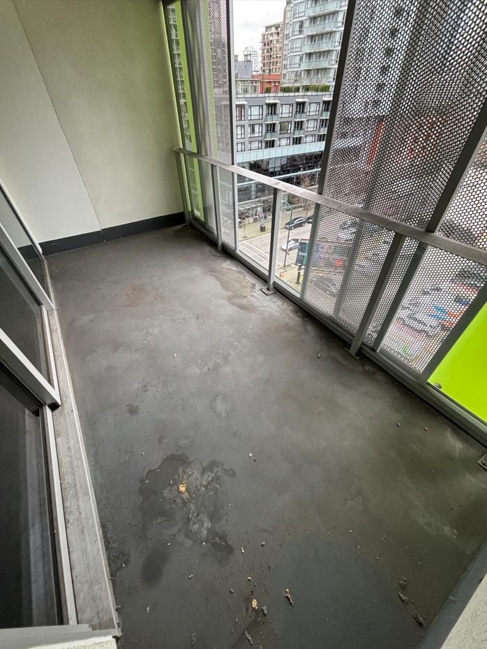 A small, partially damp balcony with concrete flooring, scattered leaves, and a glass railing overlooks a rainy, overcast street lined with trees.