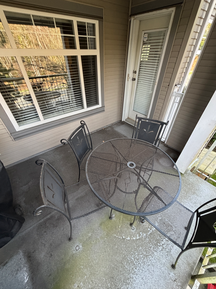 Small outdoor patio with a metal table with a glass top and four matching chairs, attached to a house with large window and a door with blinds.