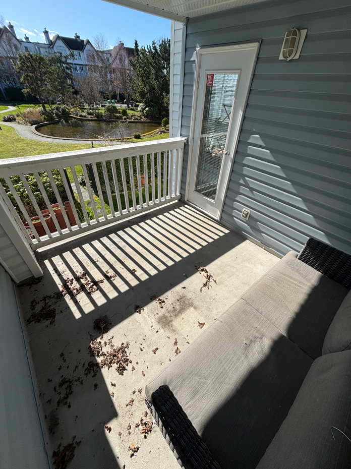 Small balcony with a beige sofa, a white railing, and a door leading inside. The floor has dried leaves and dirt, with a scenic view of a pond, trees, and houses in the background on a sunny day.