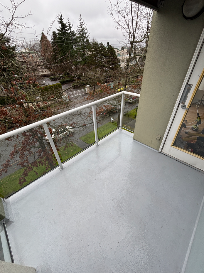 View from a small balcony with gray flooring and white railing, overlooking a tree-lined street. The scene is overcast, conveying a calm, dreary mood.