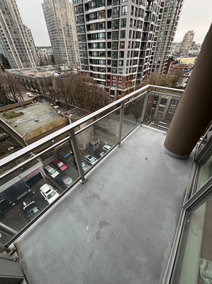 Empty balcony with glass railing, high-rise buildings in the background, parking lot below, and overcast sky.