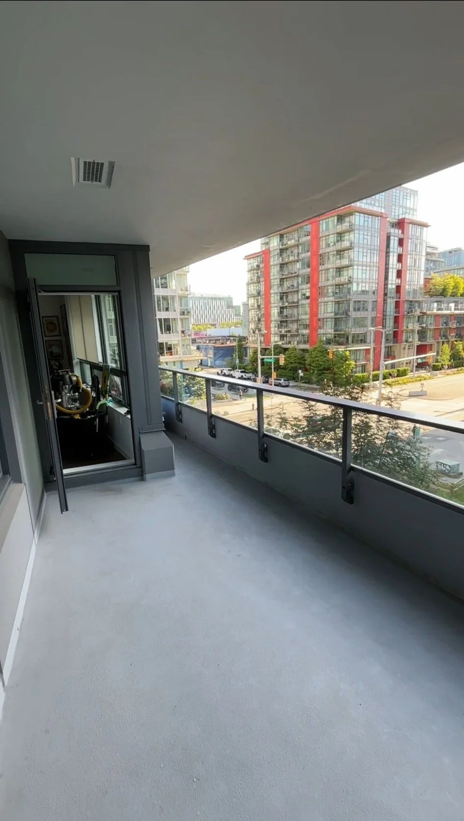 A city view from a balcony with glass railing, showing modern apartment buildings with large windows and some greenery below.