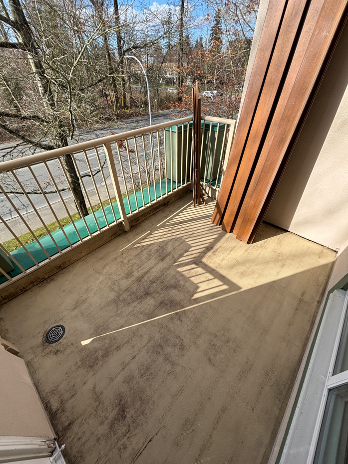 Empty balcony with a concrete floor, white metal railing, and a view of leafless trees and a street outside, with a wooden panel leaning against the wall.