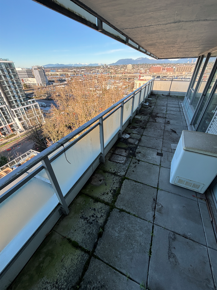 A small, partially damp balcony with concrete flooring, scattered leaves, and a glass railing overlooks a rainy, overcast street lined with trees.