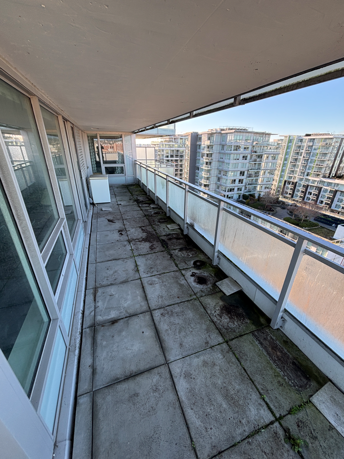 A small, partially damp balcony with concrete flooring, scattered leaves, and a glass railing overlooks a rainy, overcast street lined with trees.