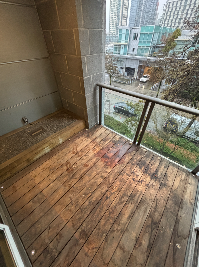 A small, partially damp balcony with concrete flooring, scattered leaves, and a glass railing overlooks a rainy, overcast street lined with trees.
