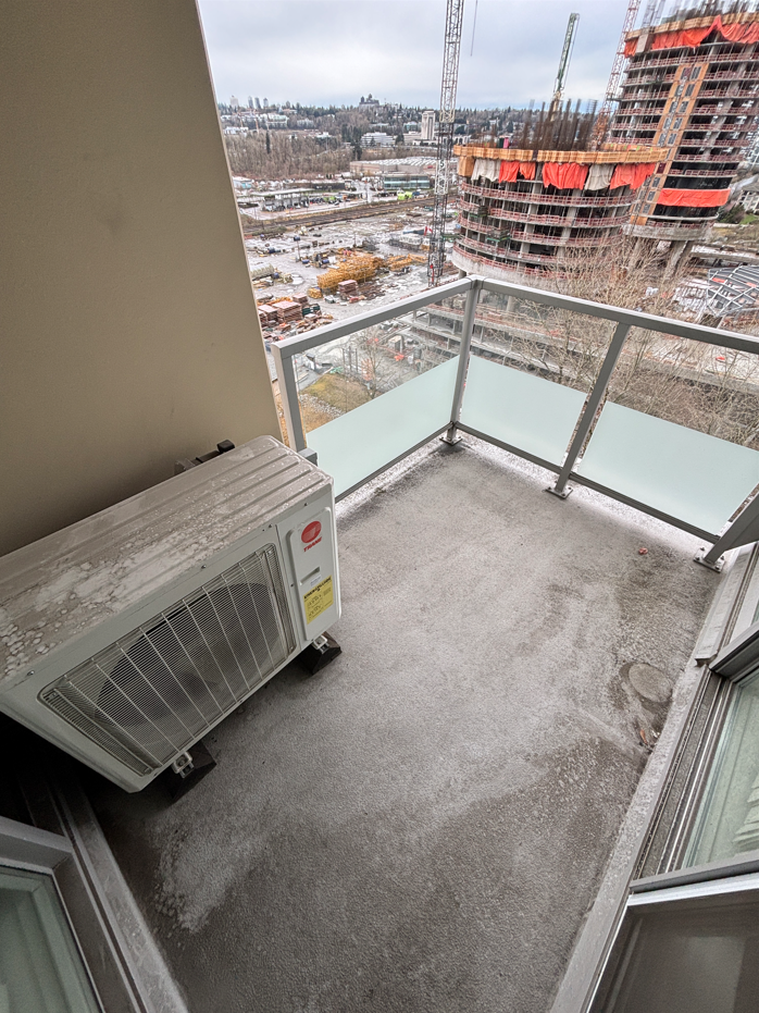 Empty balcony with concrete floor, glass railing, and an air conditioning unit, overlooking a construction site with buildings and cranes in the background.