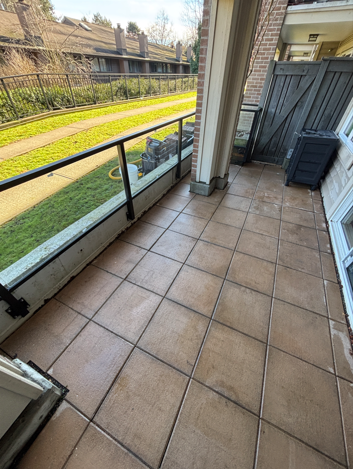 View of an apartment balcony with tiled floor, black metal railing, and outdoor storage box, overlooking a grassy area with walking path and residential buildings in the background.