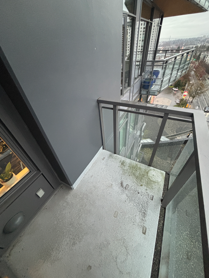 Small urban balcony with glass and metal railings, showing a city view with roads and buildings. The mood is overcast and minimalistic.