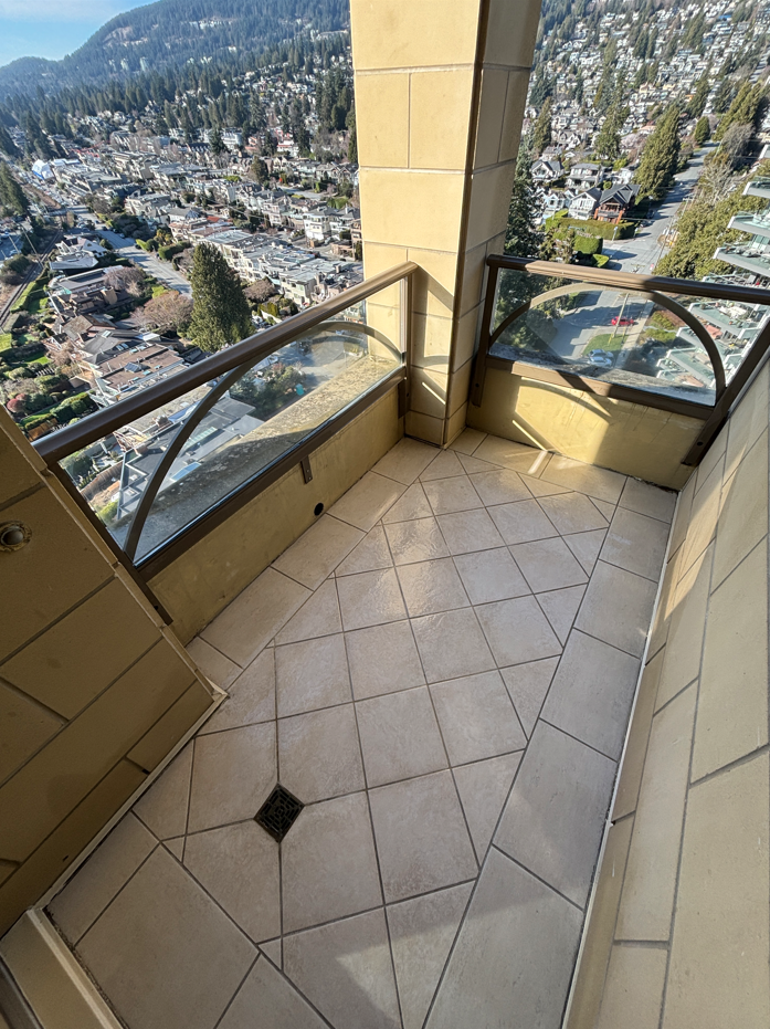 A small balcony with beige tiled flooring and glass railings, overlooking a cityscape with houses, trees, and mountains in the distance.