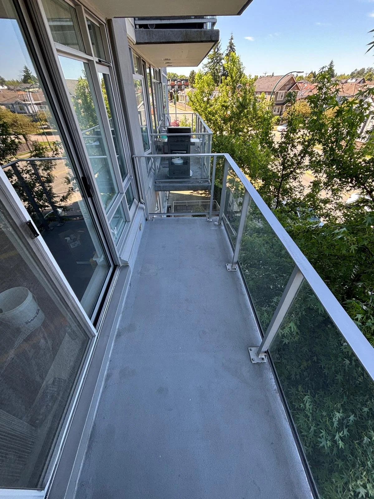 Empty balcony with glass railing, adjacent to an apartment with large windows and sliding glass doors, overlooking a tree-lined residential street with houses and trees in the background.