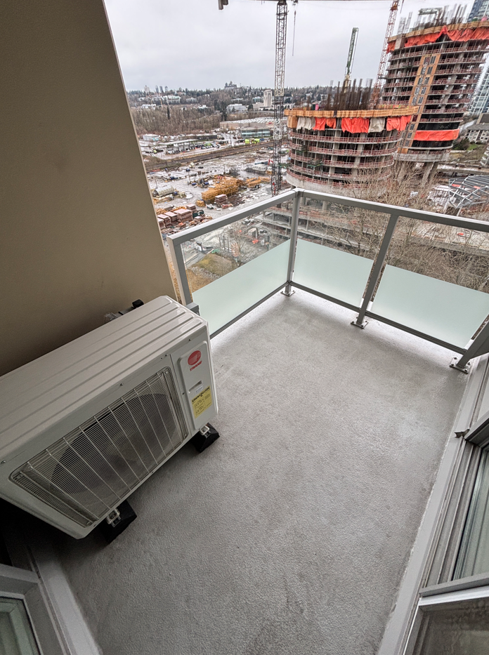 Small balcony with an air conditioning unit, frosted glass panels, and a view of a construction site with cranes and buildings under construction in the background.