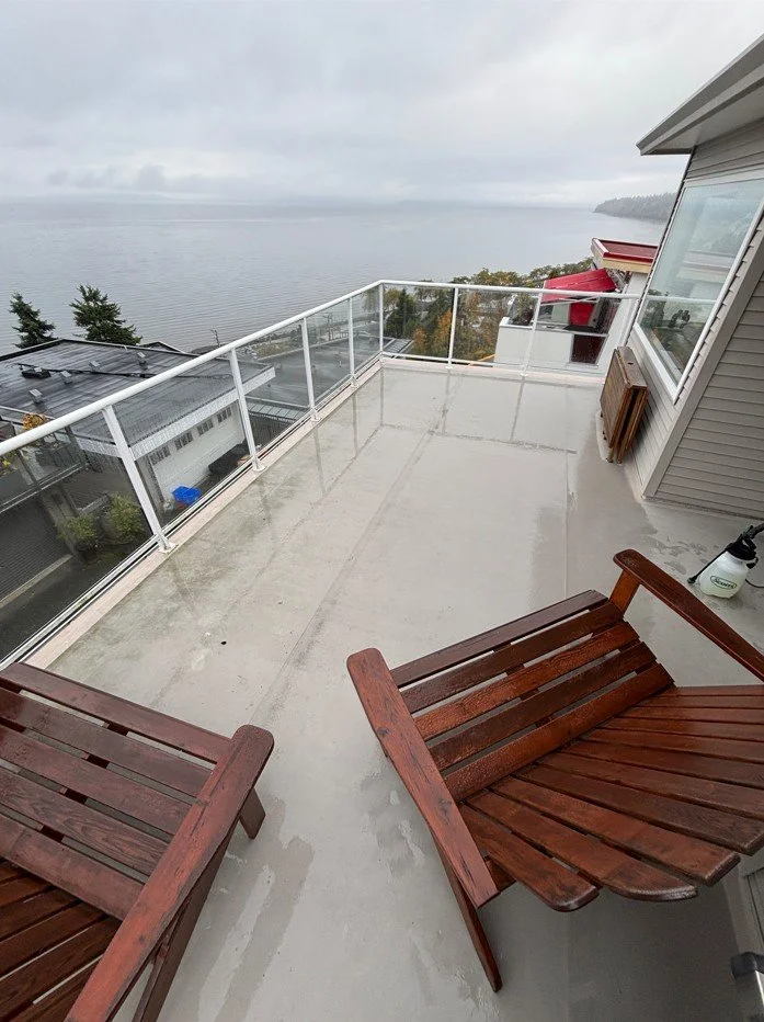 A spacious balcony with glass railing overlooking a body of water on a cloudy day. The balcony has two wooden lounge chairs, a small container near the house wall, and a red bench in the background.