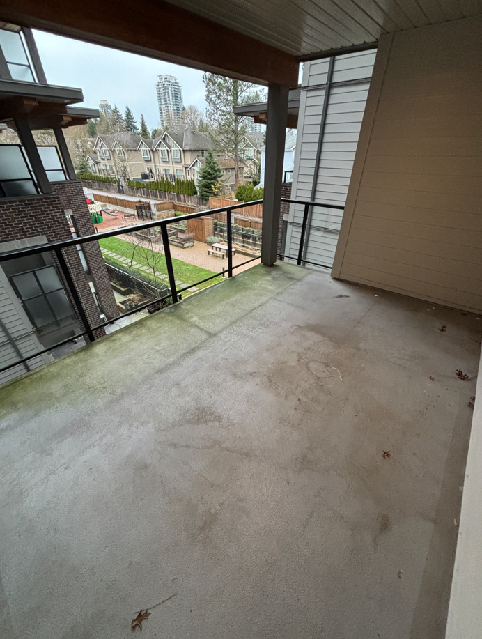 A small, partially damp balcony with concrete flooring, scattered leaves, and a glass railing overlooks a rainy, overcast street lined with trees.