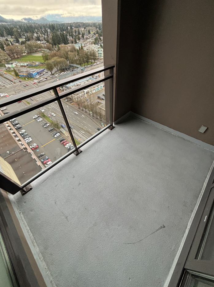 Balcony with glass railing overlooking a cityscape with parking lots, roads, a park, and mountains in the background.