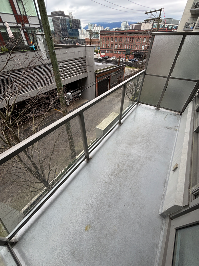 Empty balcony with glass railing overlooking a city street with buildings, trees, electrical wires, and overcast sky.