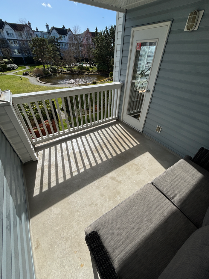 Small balcony with white railing and gray outdoor furniture, overlooking a pond and landscaped garden with trees and multi-story residential buildings in the background.