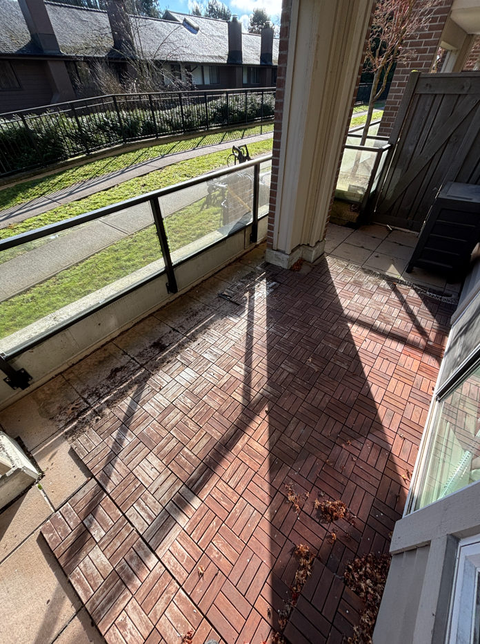 A small balcony with a brick floor, glass railing, and a view of a grass lawn and neighboring houses.