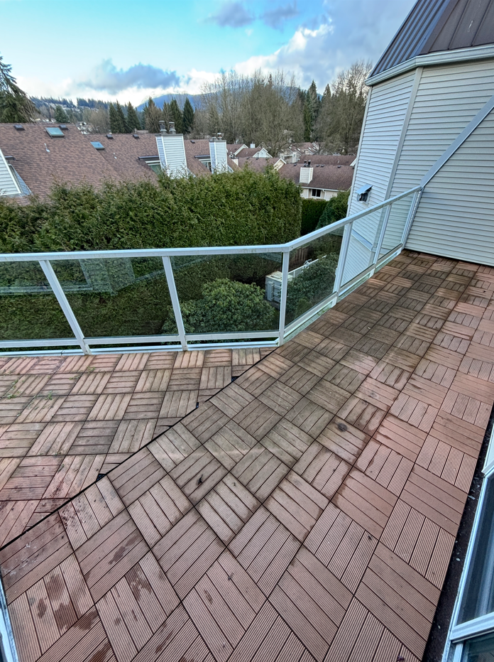 A wooden balcony with a glass railing overlooking houses with brown roofs and a background of trees and hills.
