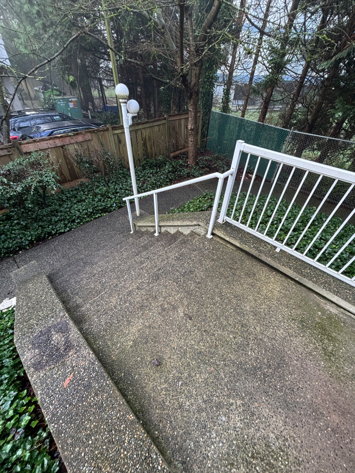 A small, partially damp balcony with concrete flooring, scattered leaves, and a glass railing overlooks a rainy, overcast street lined with trees.