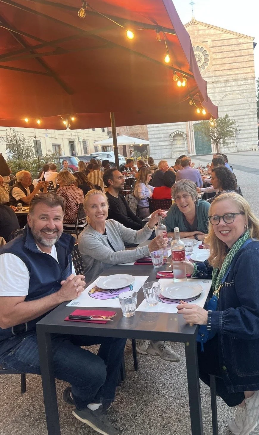People enjoying dinner at an outdoor restaurant under string lights, with a historic building in the background.