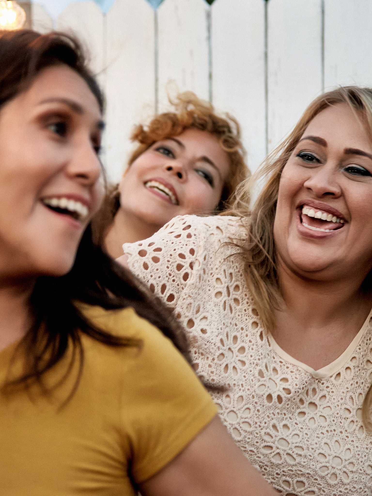 Close-up of three women smiling and laughing, with two women in the foreground and one in the background, at a social gathering.