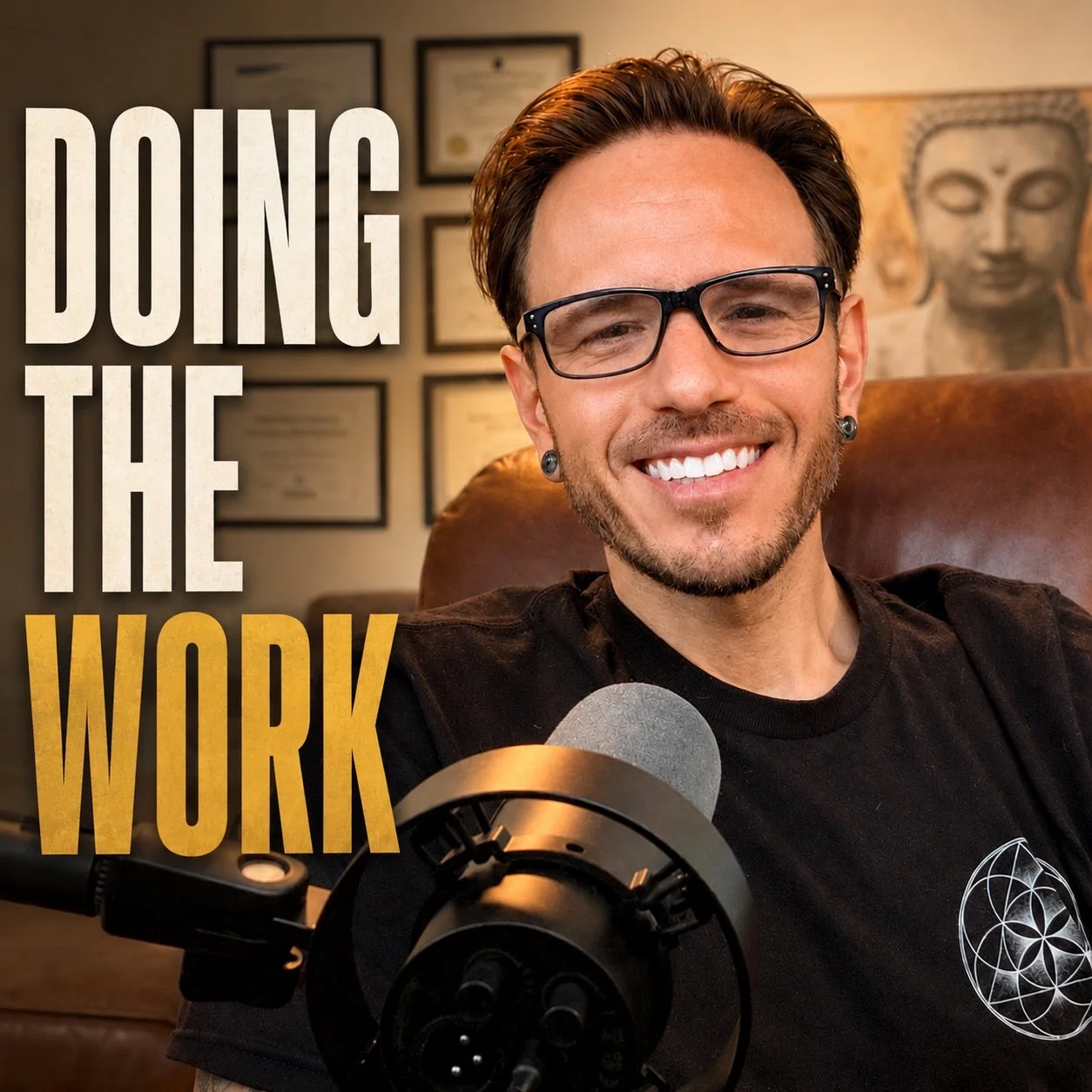 A man with glasses and earrings smiling at the camera, sitting in front of a microphone, with framed certificates on the wall and a Buddha artwork in the background, in a home office.