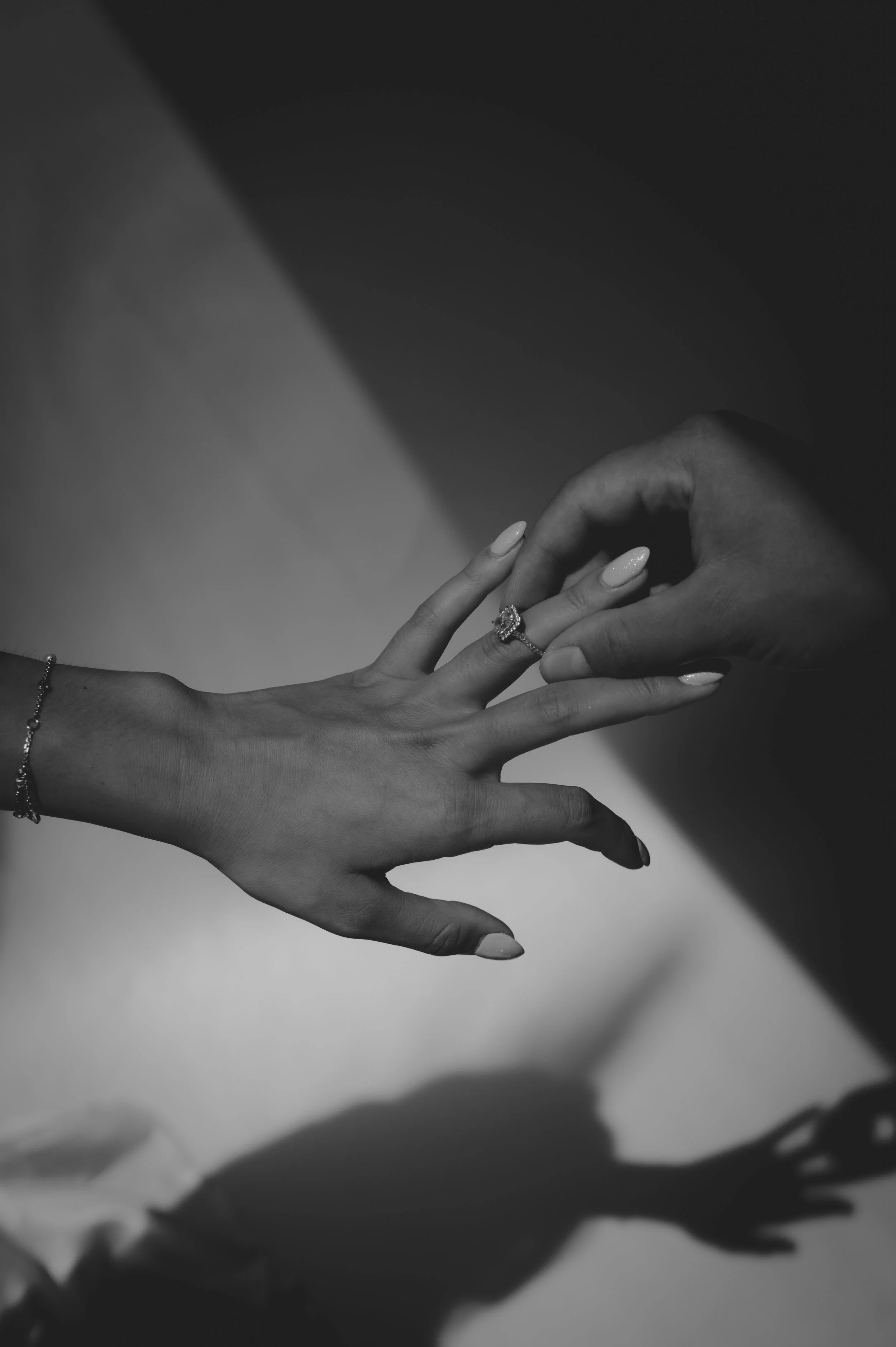Two hands are reaching toward each other, fingers slightly touching, with one hand resting on a surface and the other hand with a ring on the ring finger. The image is in black and white.