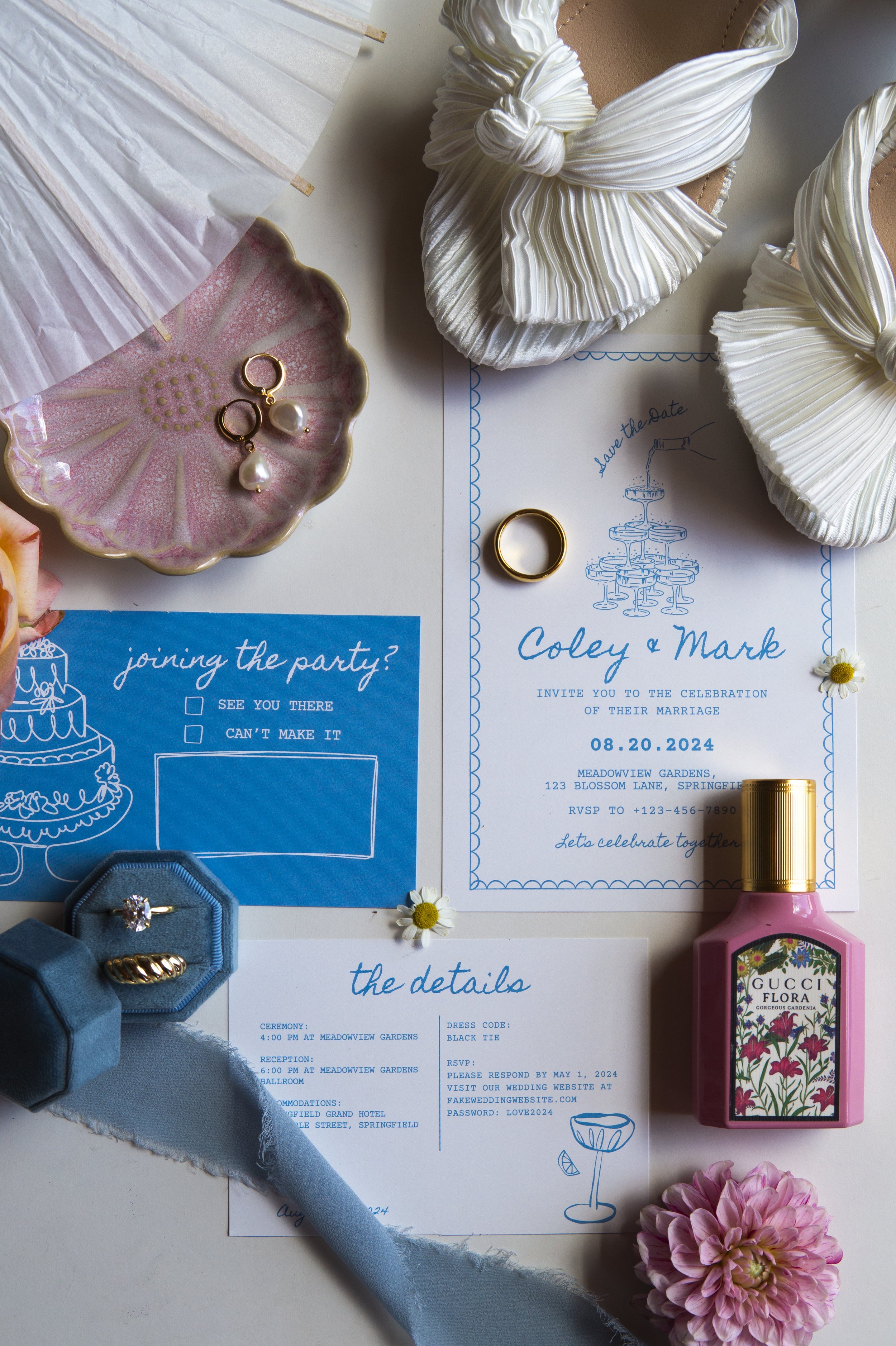 Wedding invitation surrounded by a pair of satin shoes with bows, a pink framed bottle of Gucci Flora perfume, a pink flower, and a blue ribbon, along with a pink ceramic dish holding pearl and gold earrings, a gold ring, and floral decorations.