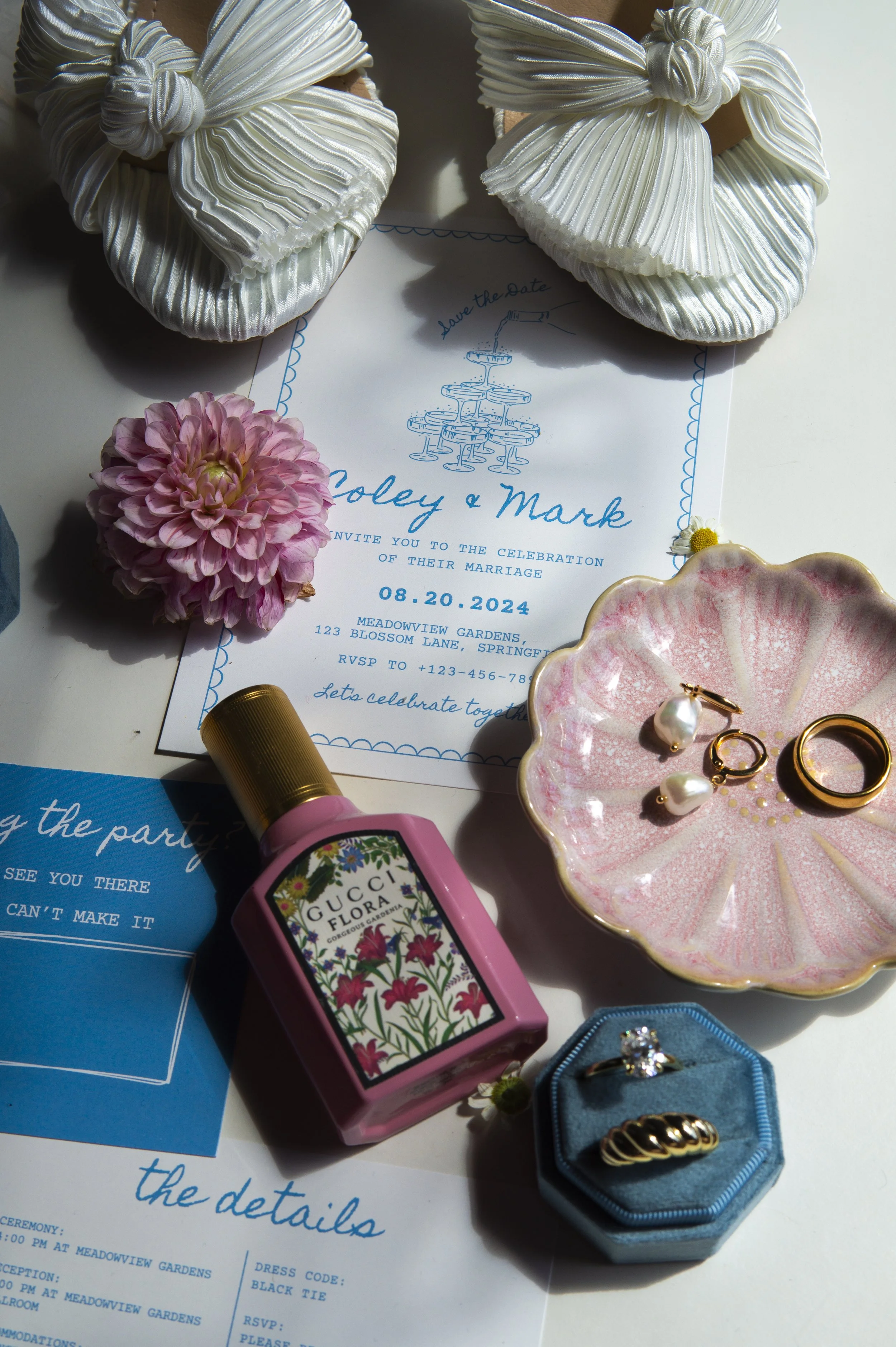 A wedding invitation surrounded by various accessories including a pink flower, a ceramic dish with jewelry, a lipstick, a small blue box, and several rings, with two white satin slippers at the top of the image.