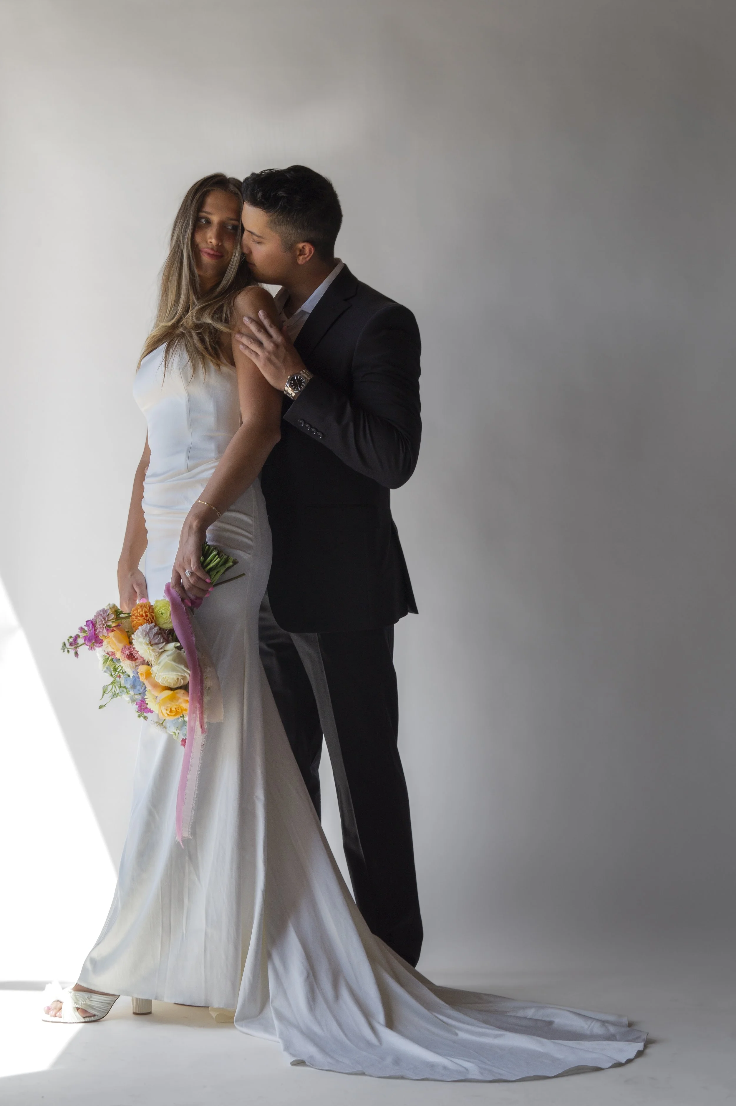 A bride in a white gown holding a bouquet of colorful flowers stands close to a groom in a black tuxedo, both gazing at each other in an indoor setting with a plain white background.