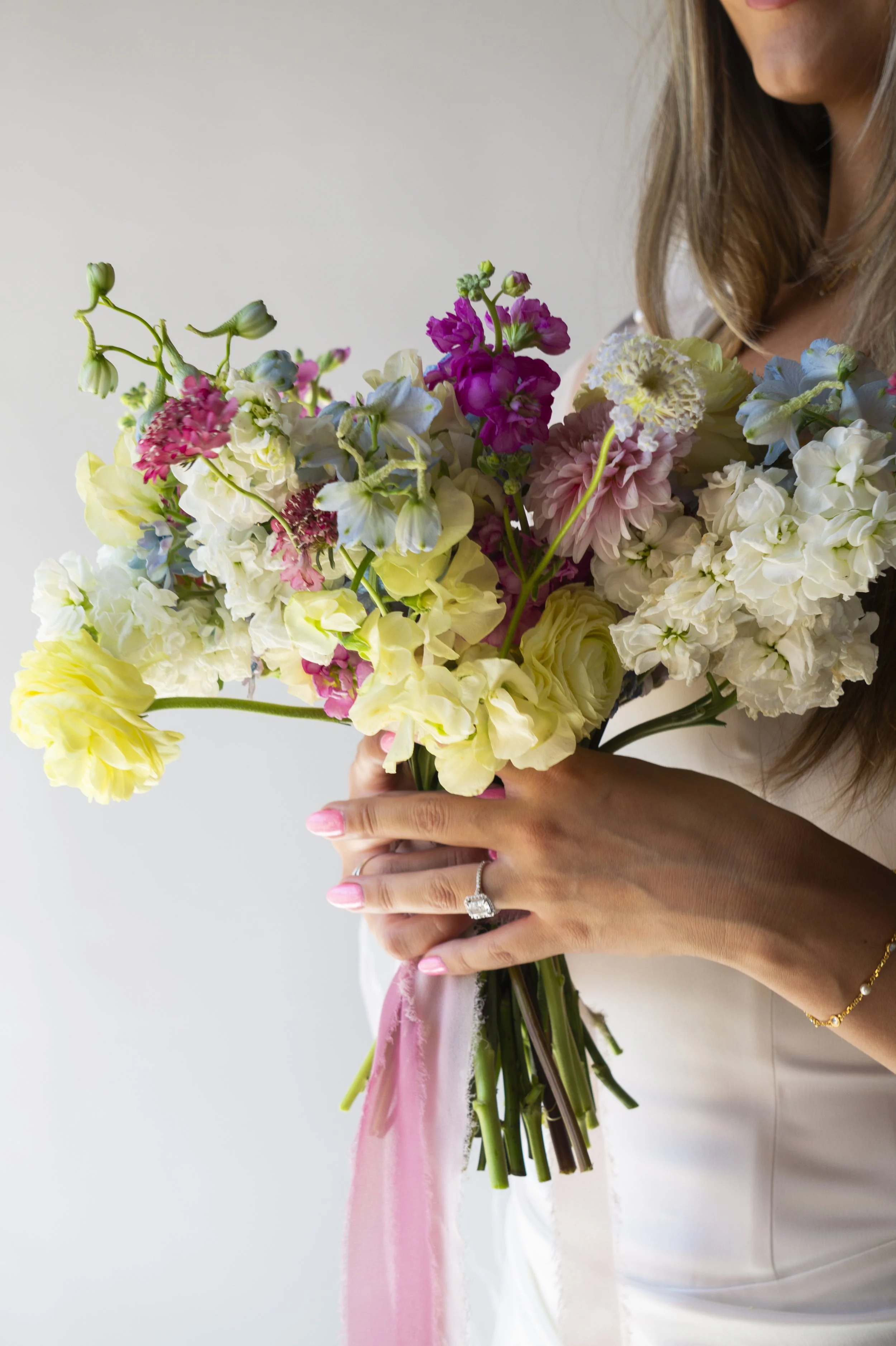 Person holding a bouquet of colorful flowers, wearing a ring on her finger, a gold bracelet, and a white dress, with a soft smile and long hair.