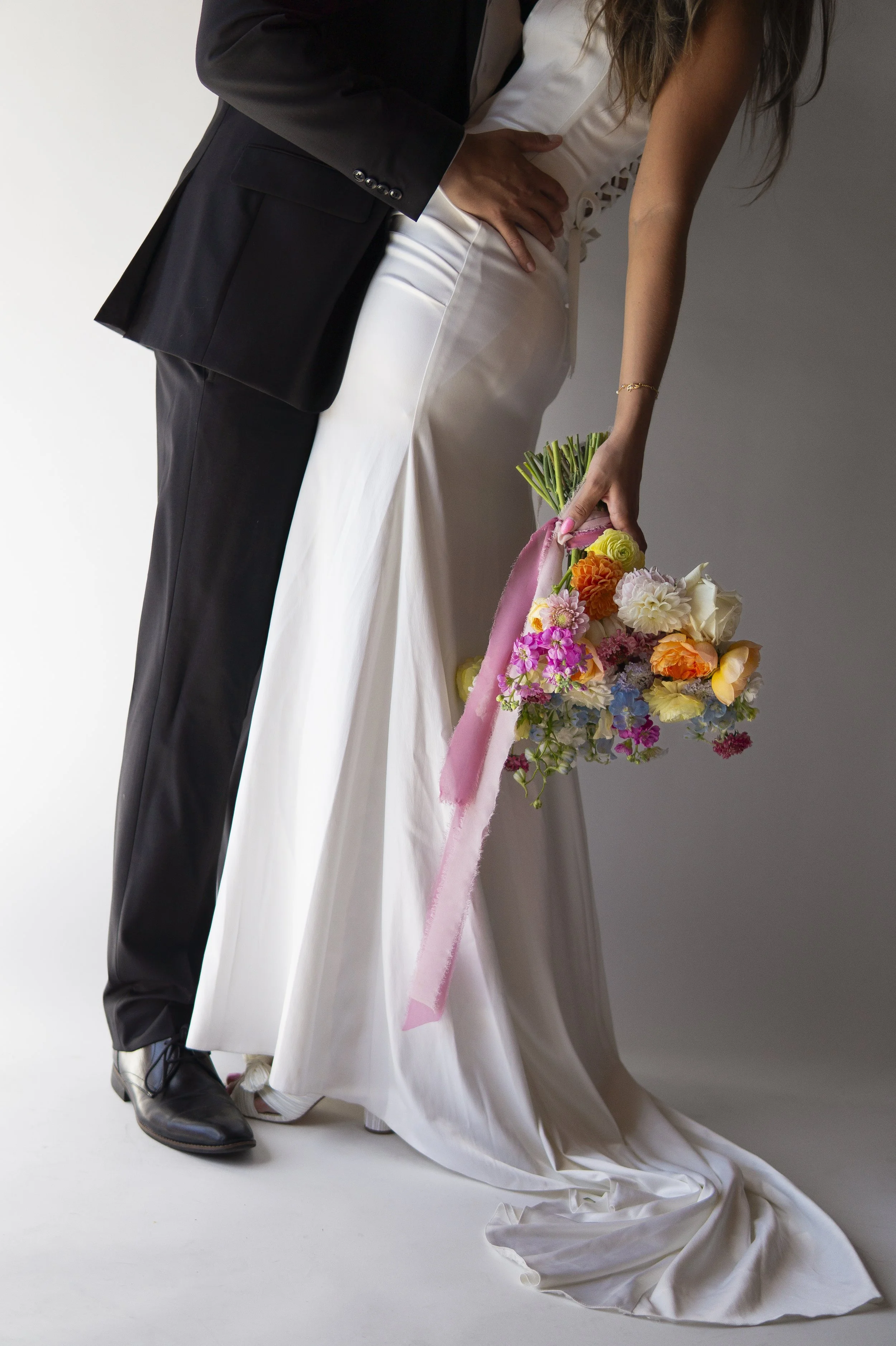 A couple in wedding attire, with the man in a black suit and the woman in a white wedding dress, holding a bouquet of colorful flowers on a white backdrop.