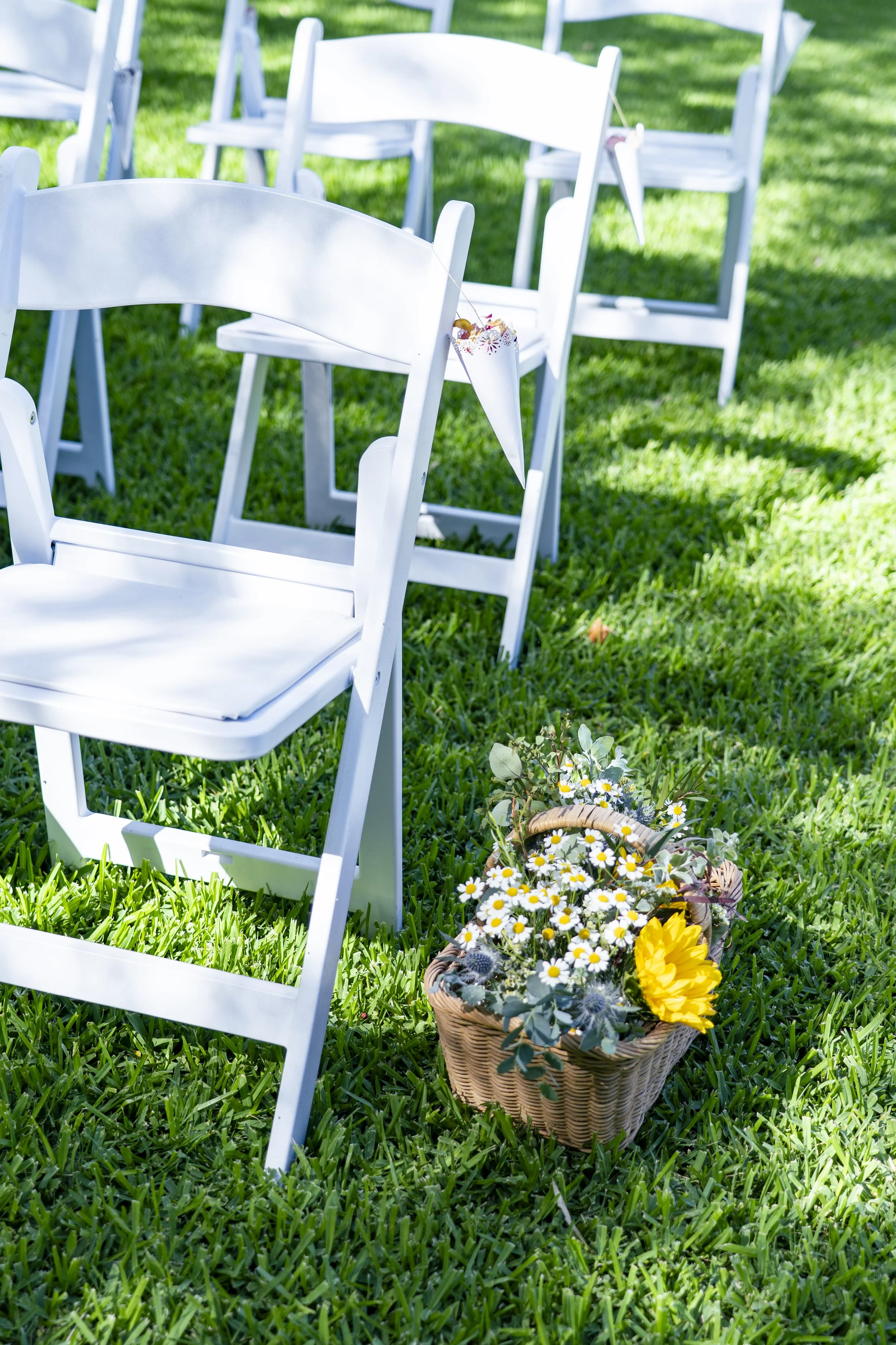 White chairs arranged outdoors on green grass, with a basket of colorful flowers in the foreground.
