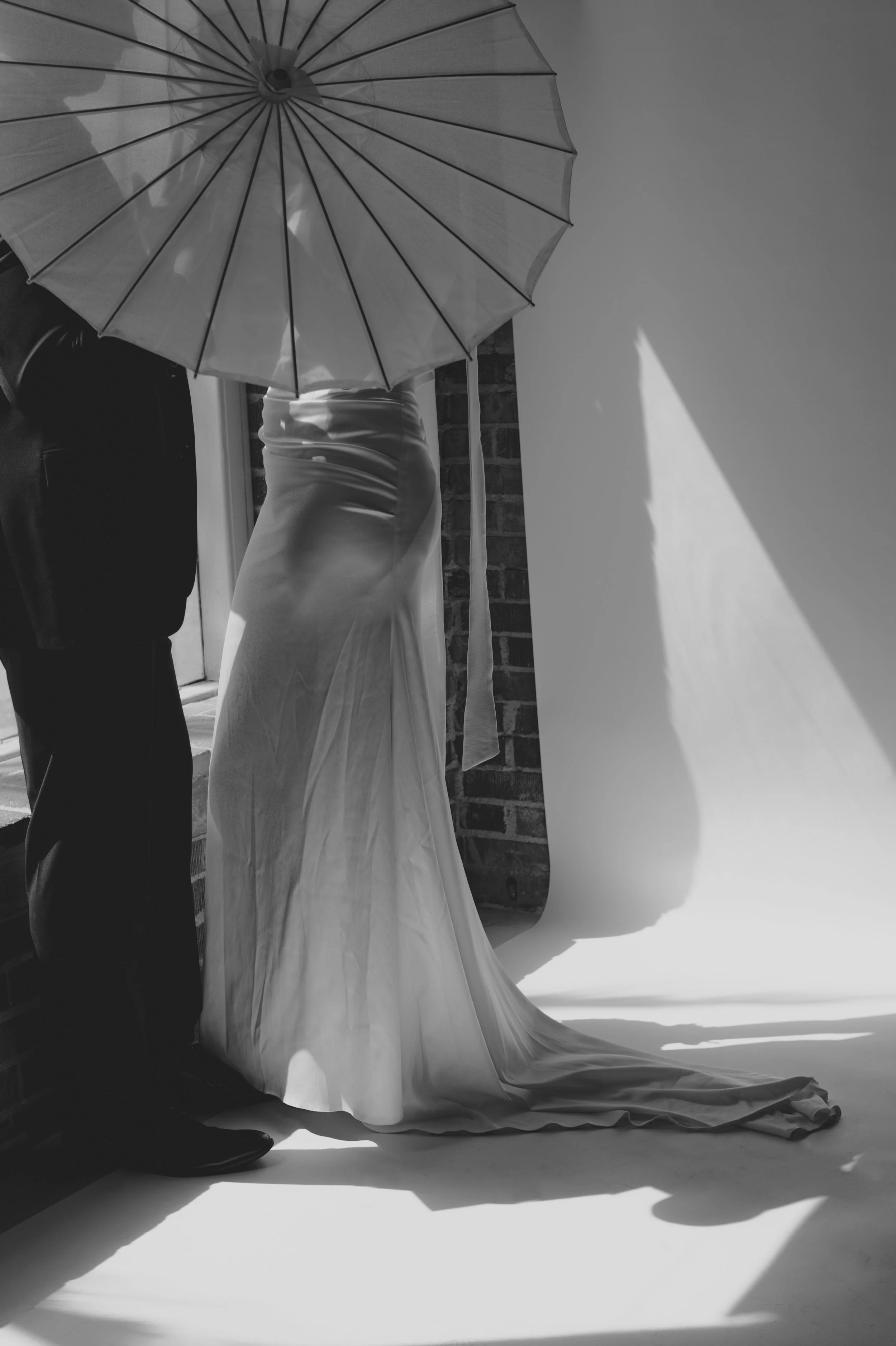 A black and white photo of a bride in a wedding dress with a fitted waist and flowing train, partial view of a man in a suit holding a large open umbrella, set against a white backdrop with shadows cast on the wall.