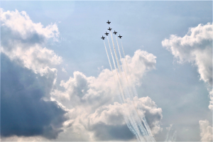 A formation of planes flying in a V-shape, leaving white smoke trails in a blue sky with clouds.