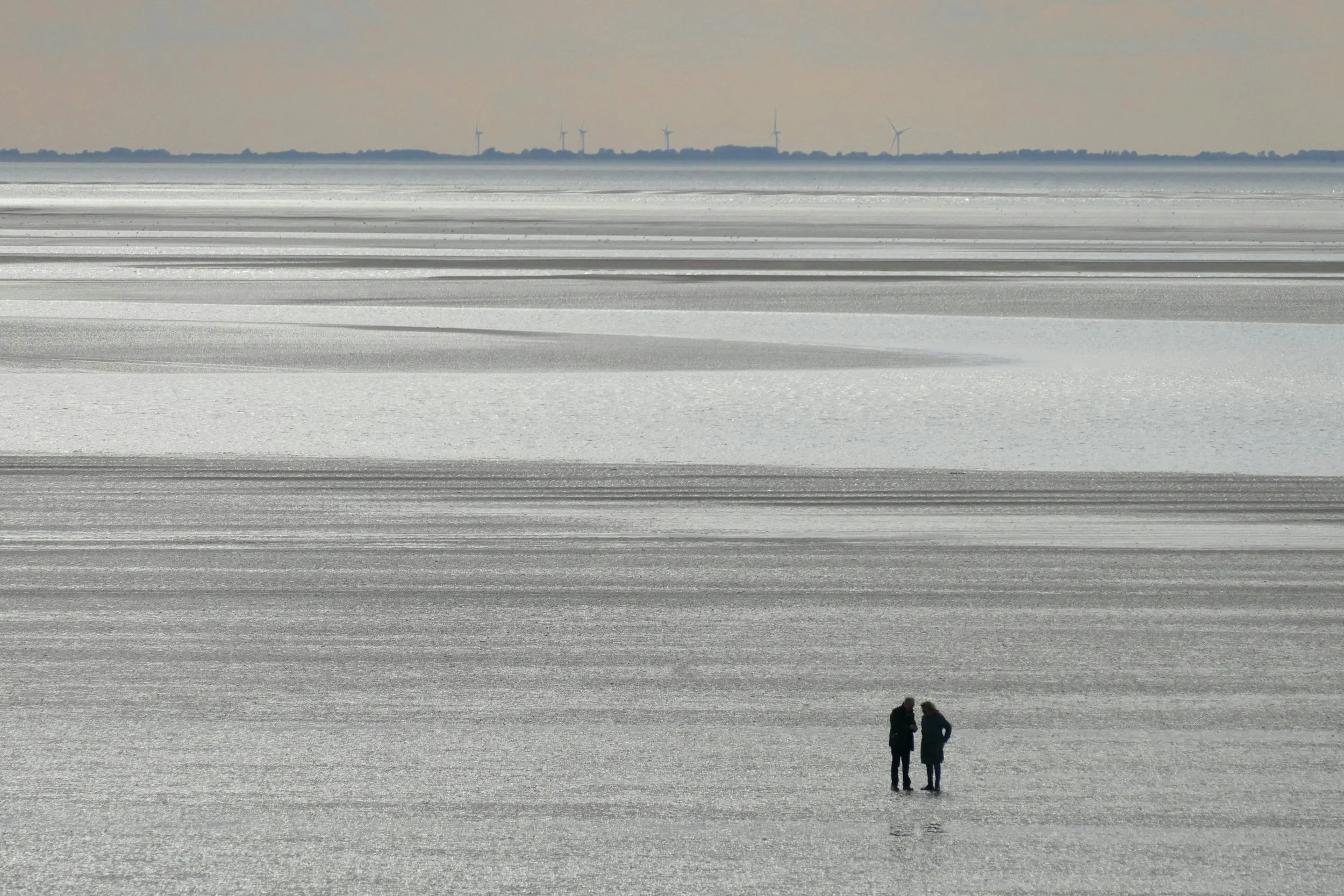 Two people standing on a snowy, reflective landscape with wind turbines visible in the distance.