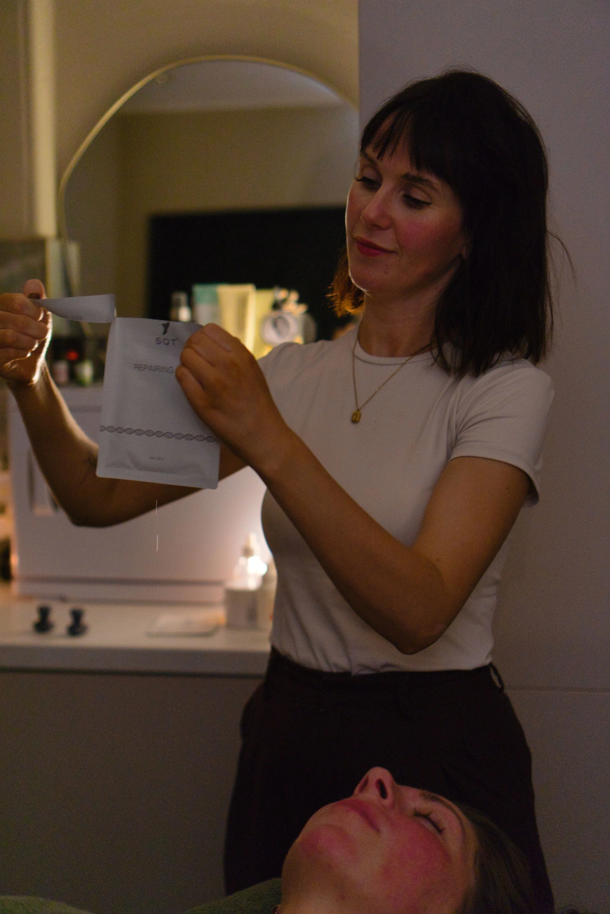 A woman with dark hair and a white shirt is holding a small paper bag labeled 'SOT' and opening it, standing beside a woman lying down with her eyes closed. The setting appears to be a cozy living room or kitchen with warm lighting.