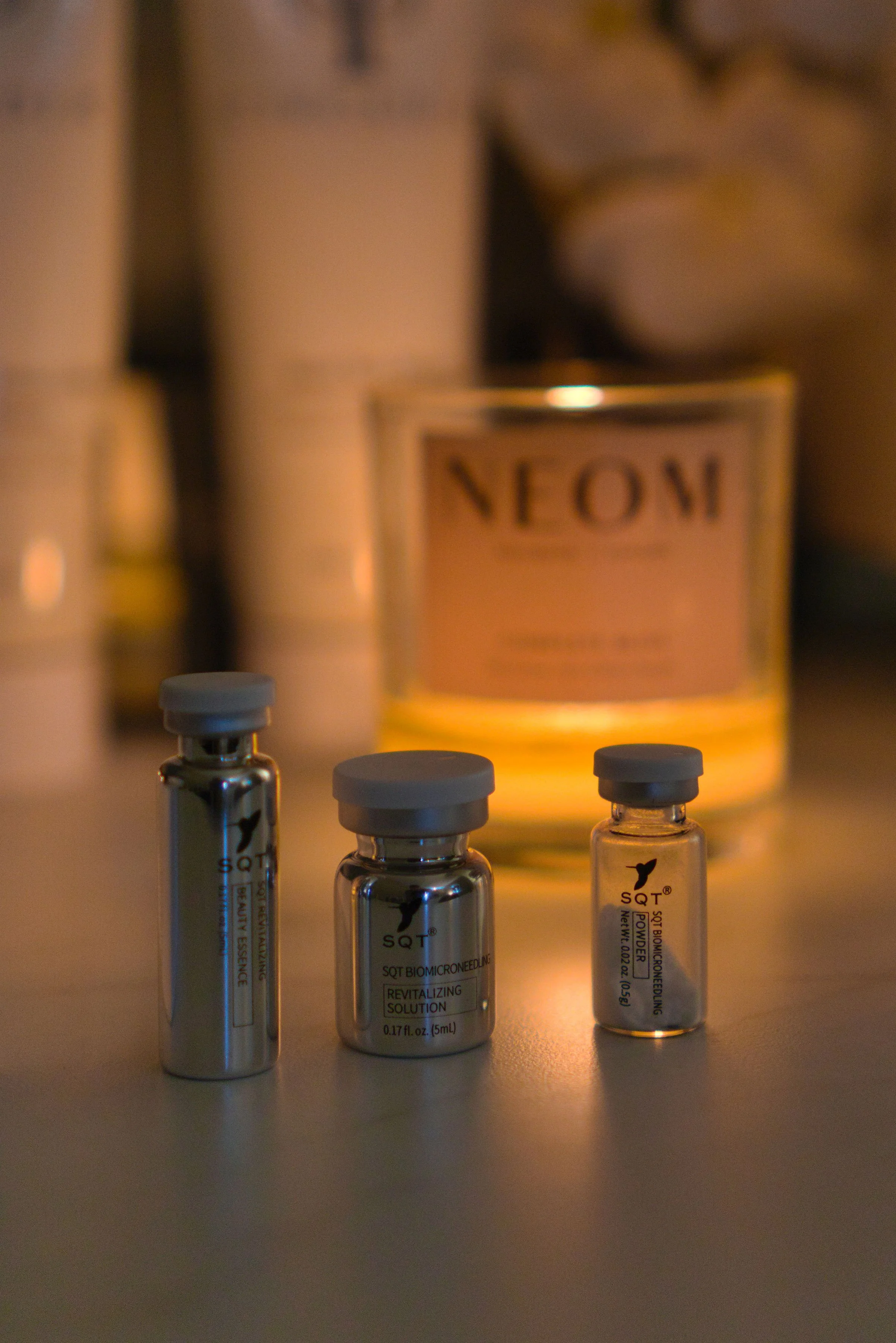 Three small metal vials with white caps on a counter in front of a lit candle labeled 'NEOM,' with blurred background of additional containers.