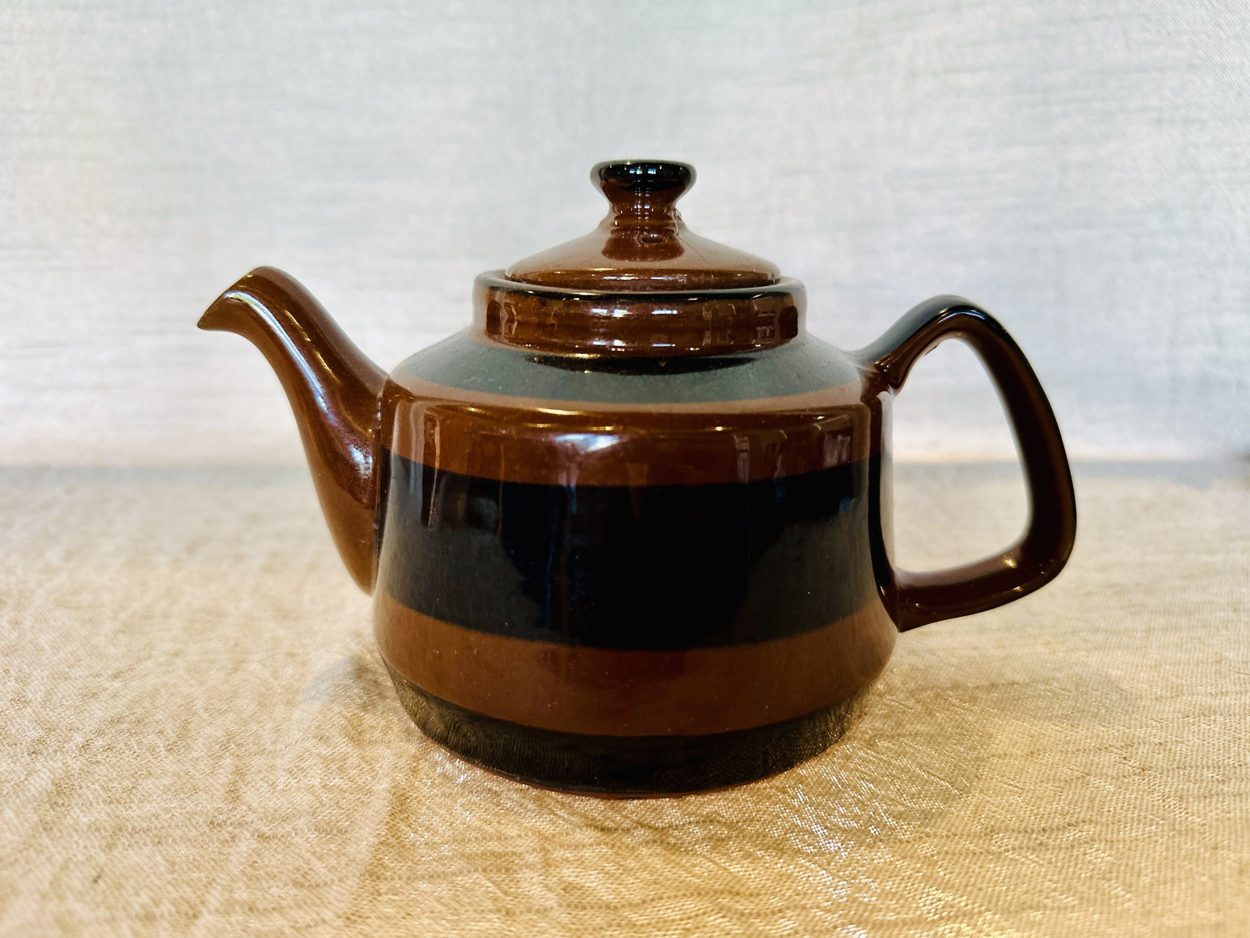 A ceramic teapot with a brown and black striped pattern, a round lid, a curved spout, and an angular handle, placed on a beige surface. Available in the Curated by Curtis online Vintage Store. 