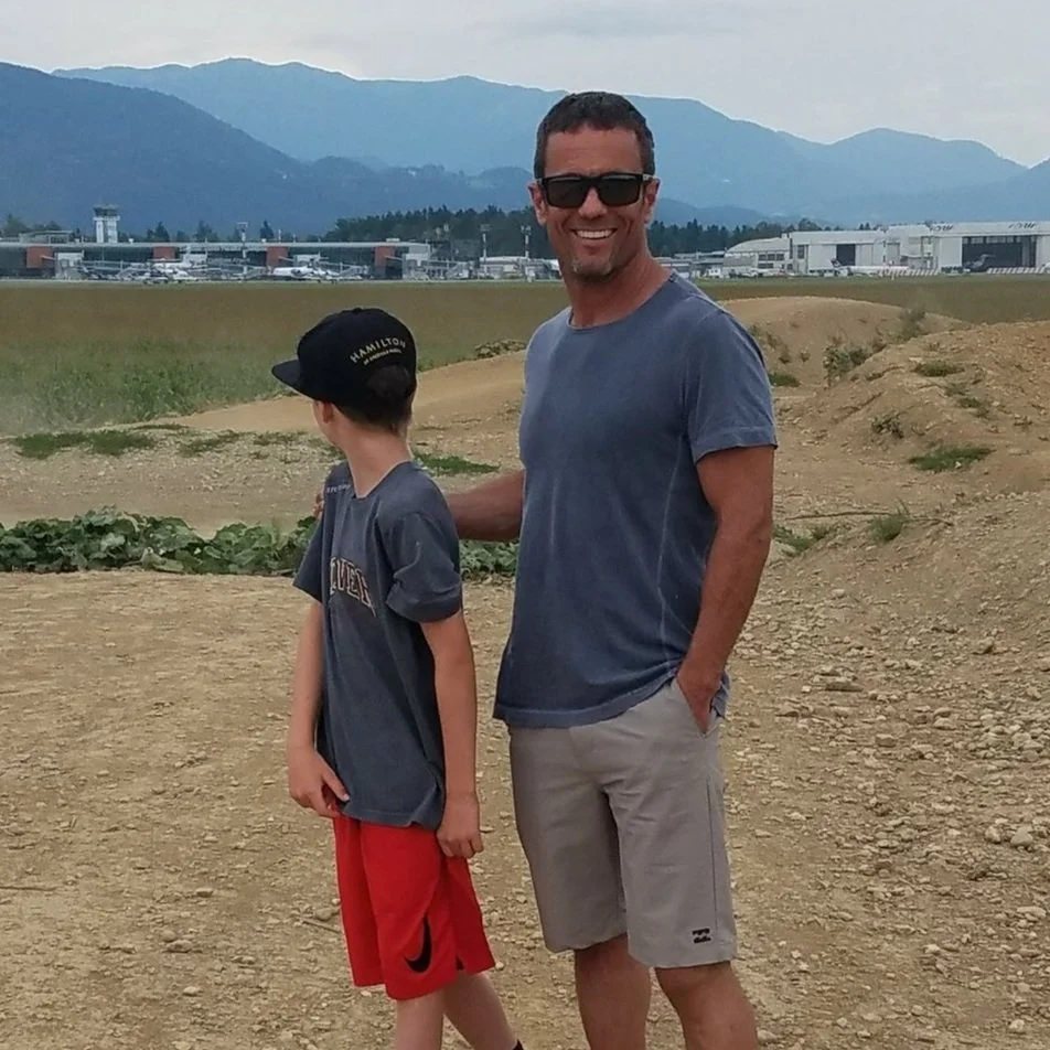 Man in sunglasses with child wearing a cap, standing outdoors on a dirt path, with mountains and an airport in the background.