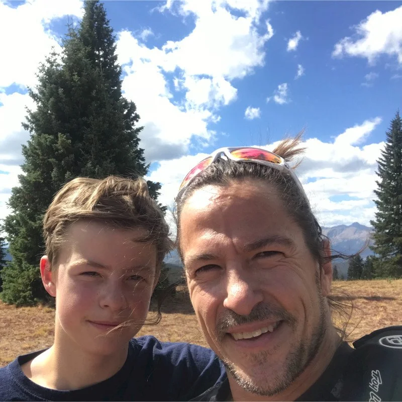 Two people outdoors in a scenic mountain landscape with pine trees and a blue sky with clouds.