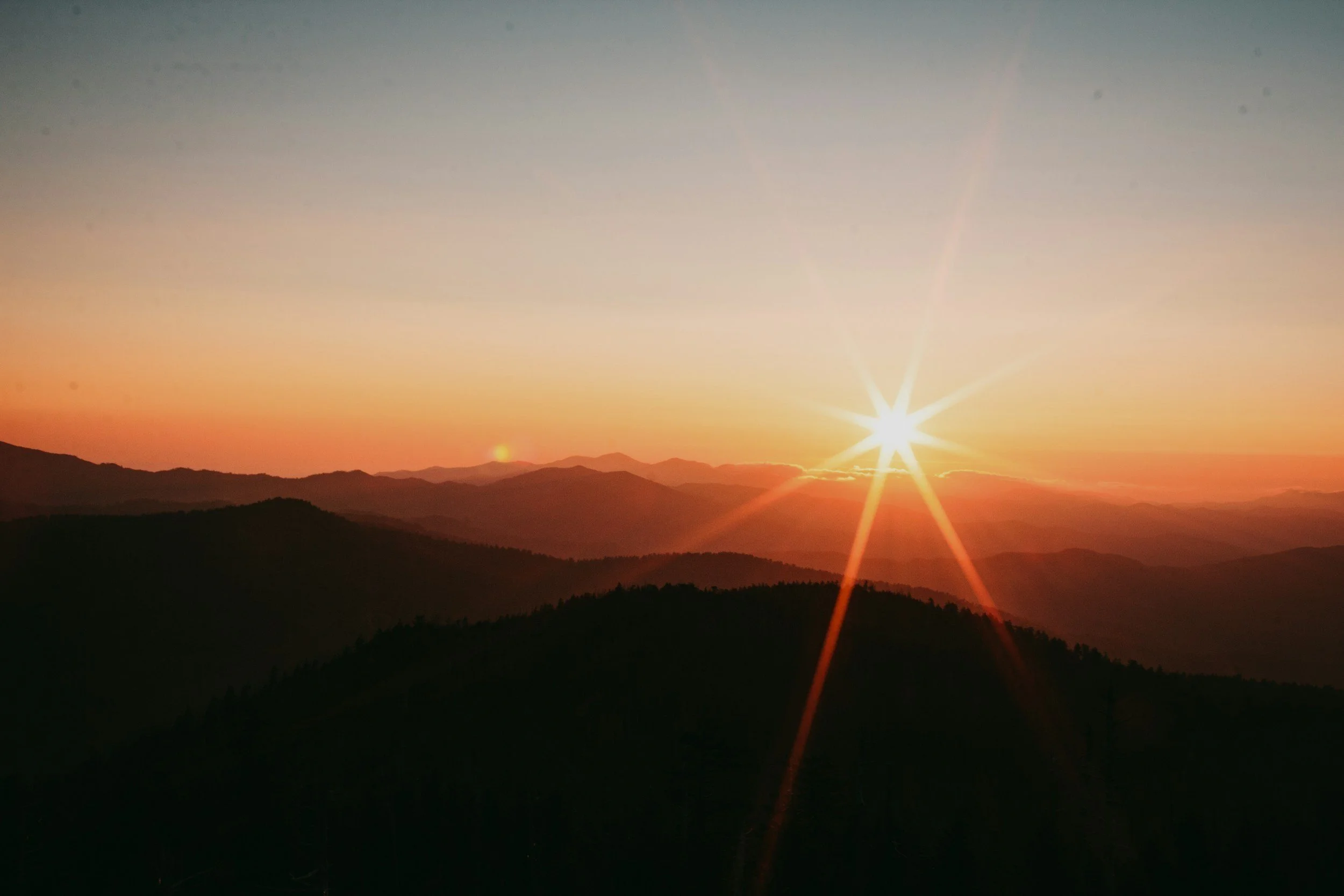 Sunset over a mountain range with a radiant sun near the horizon and a pink-orange sky.