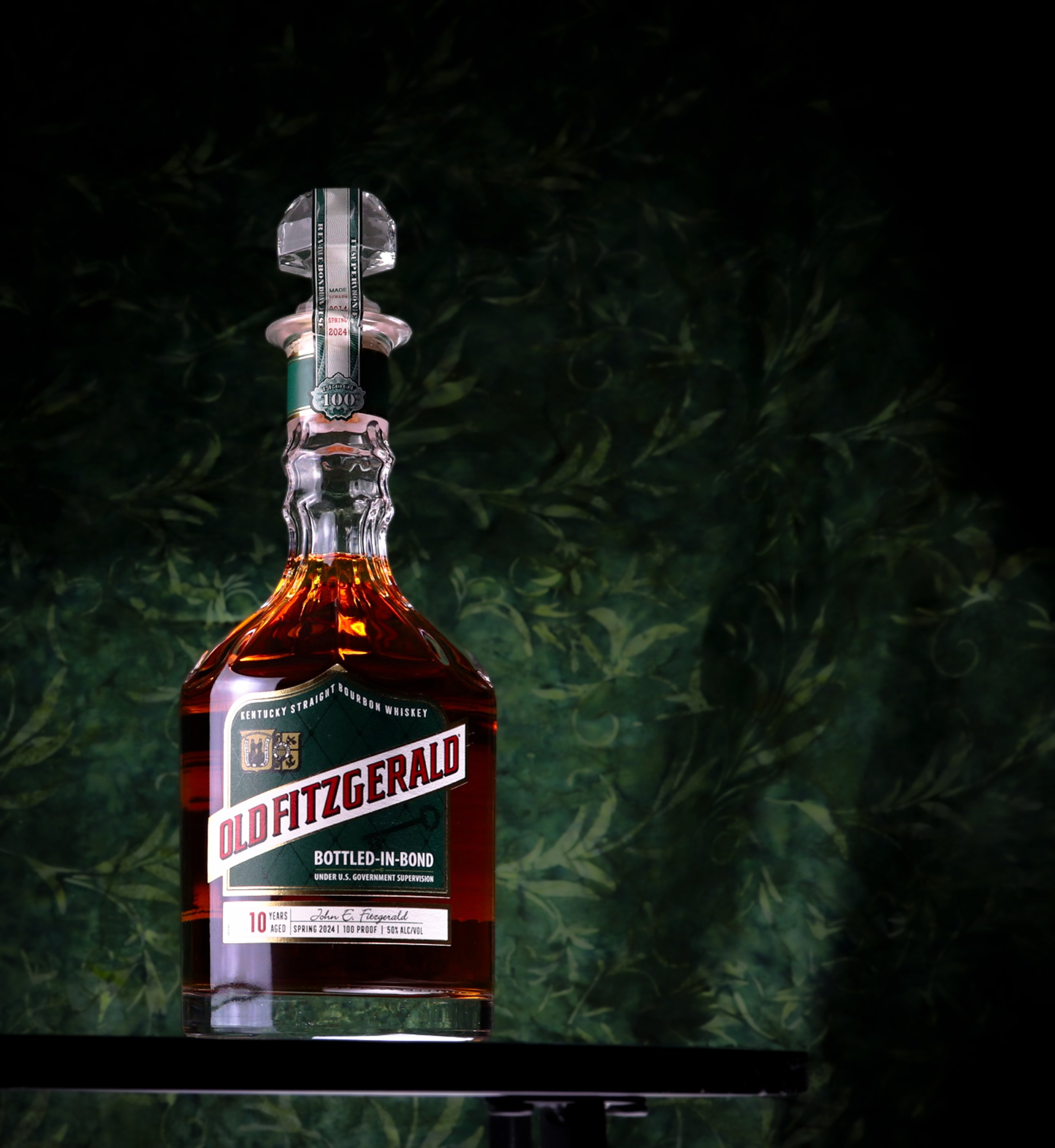 A bottle of Old Fitzgerald bourbon whiskey with a decorative decanter stopper, standing on a black surface against a dark leafy background.