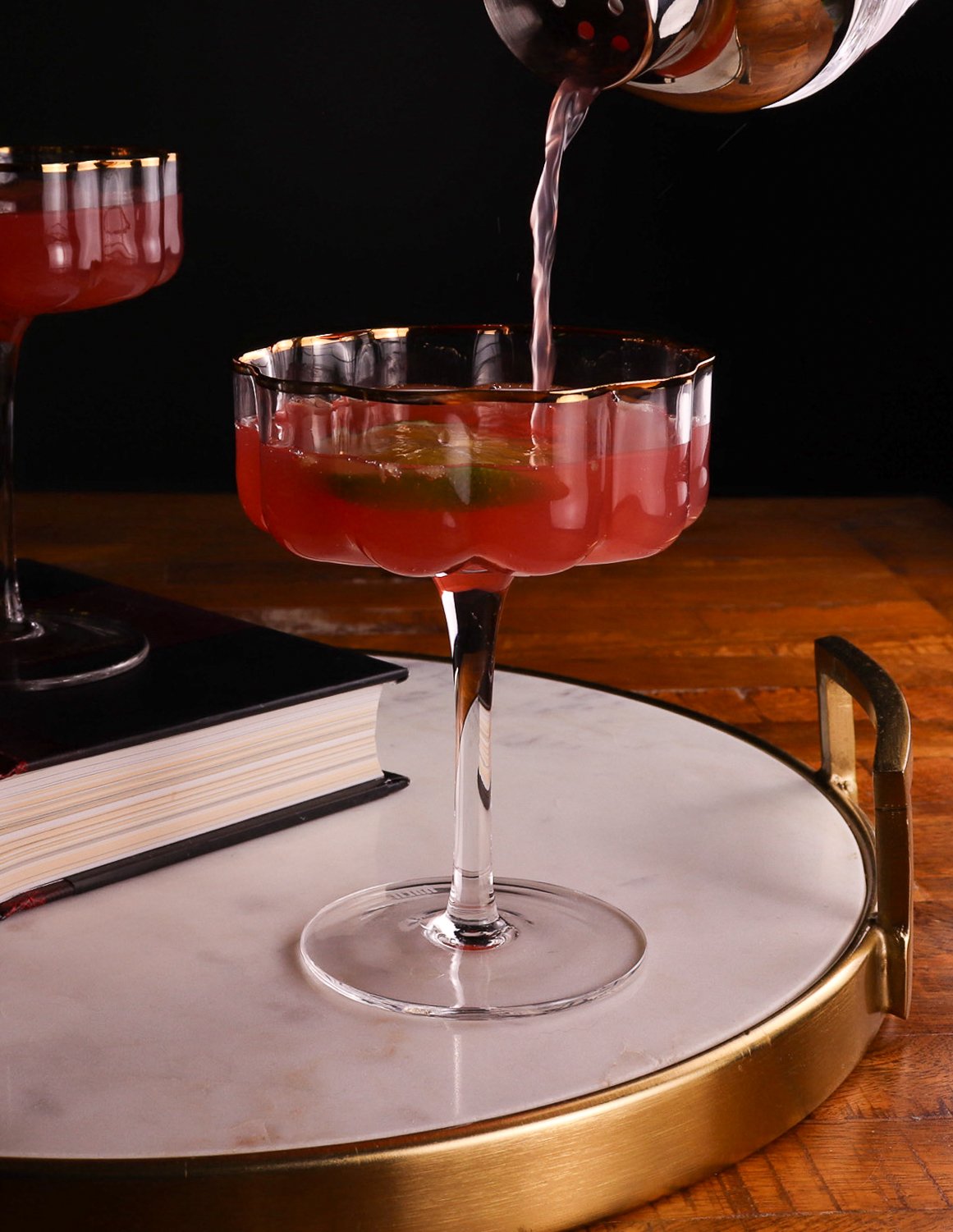 A pink cocktail in a decorative glass with a slice of lime, being poured from a cocktail shaker, set on a round tray with a book nearby.